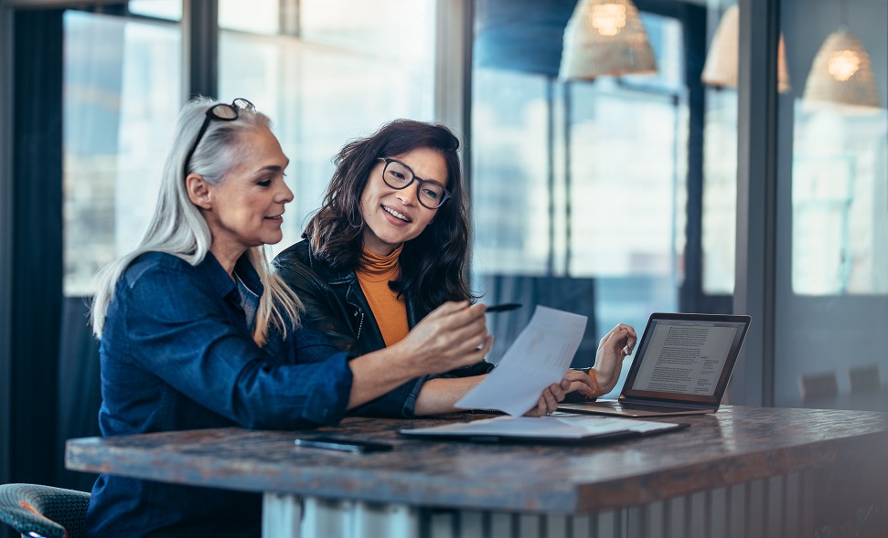 Two female professionals appearing to be having a work discussion with a laptop and paperwork at a desk