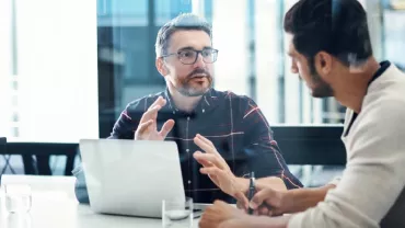 Two men having a discussion in an office with a laptop open on the table.