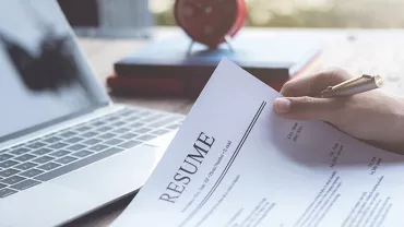 Person holding a resume in front of a laptop on a wooden desk.