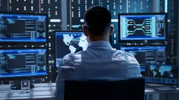 Man in front of multiple screens showing data in a dark server room.