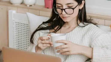 Woman with glasses holding a mug and using a laptop in a cozy room.