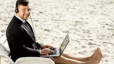 Man in a suit using a laptop and headset on the beach.